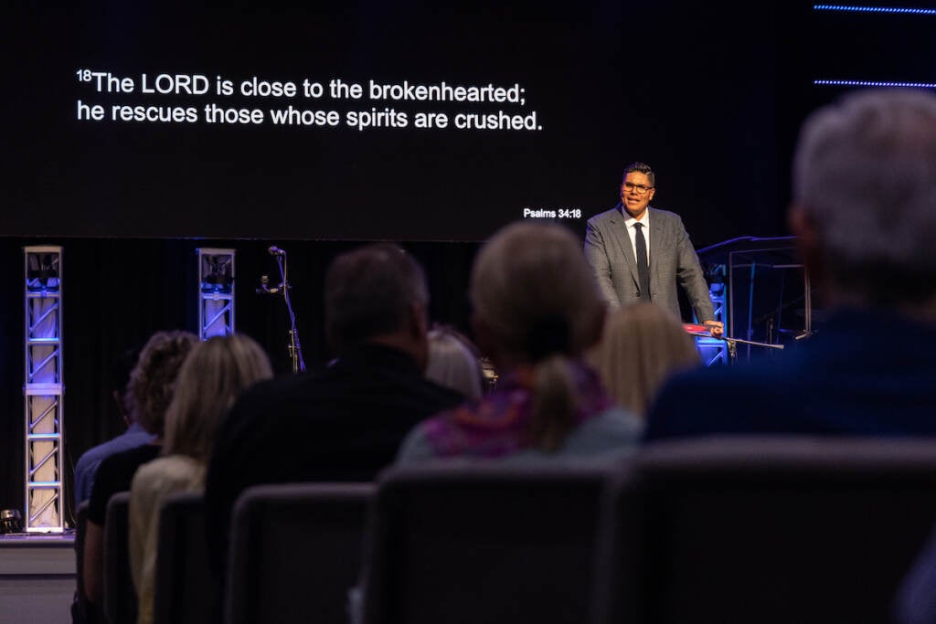 UMC Pastor George Matthew Clash speaks in front of people at a vigil. A quote from Psalms is displayed on the screen behind him, reading: "The Lord is close to the brokenhearted; he rescues those whose spirits are crushed."