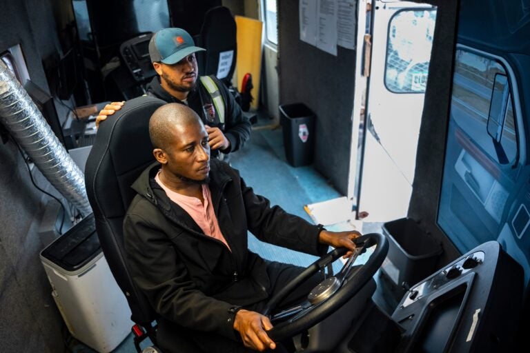 Trucking student Mohammed Kamal, left, overseen by instructor Tom Panton, practices driving a tractor-trailer truck in a simulation